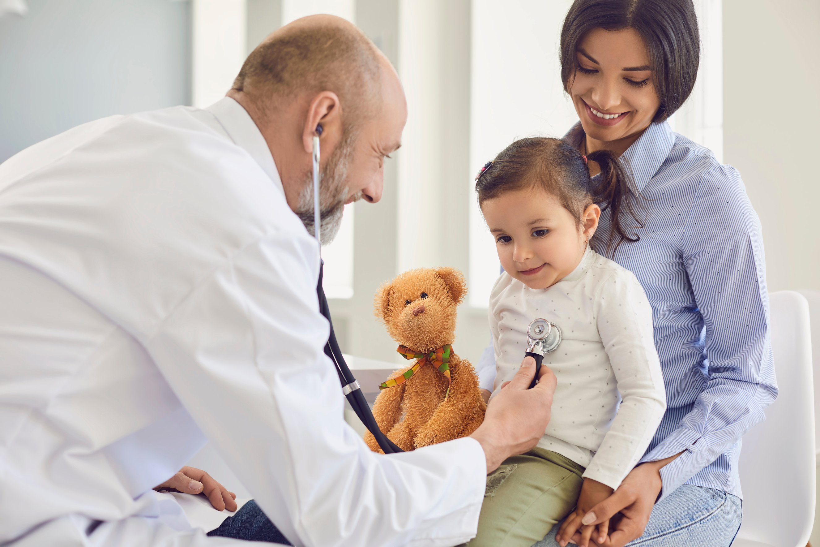 Family with a Visit to the Doctor in the Clinic Office. Senior Doctor with a Stethoscope Listens to a Little Girl with Mother Patients in the Hospital.